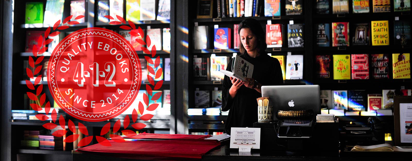 Picture of a bookseller, standing behind the counter, reading a book. There are shelves of books behind her.